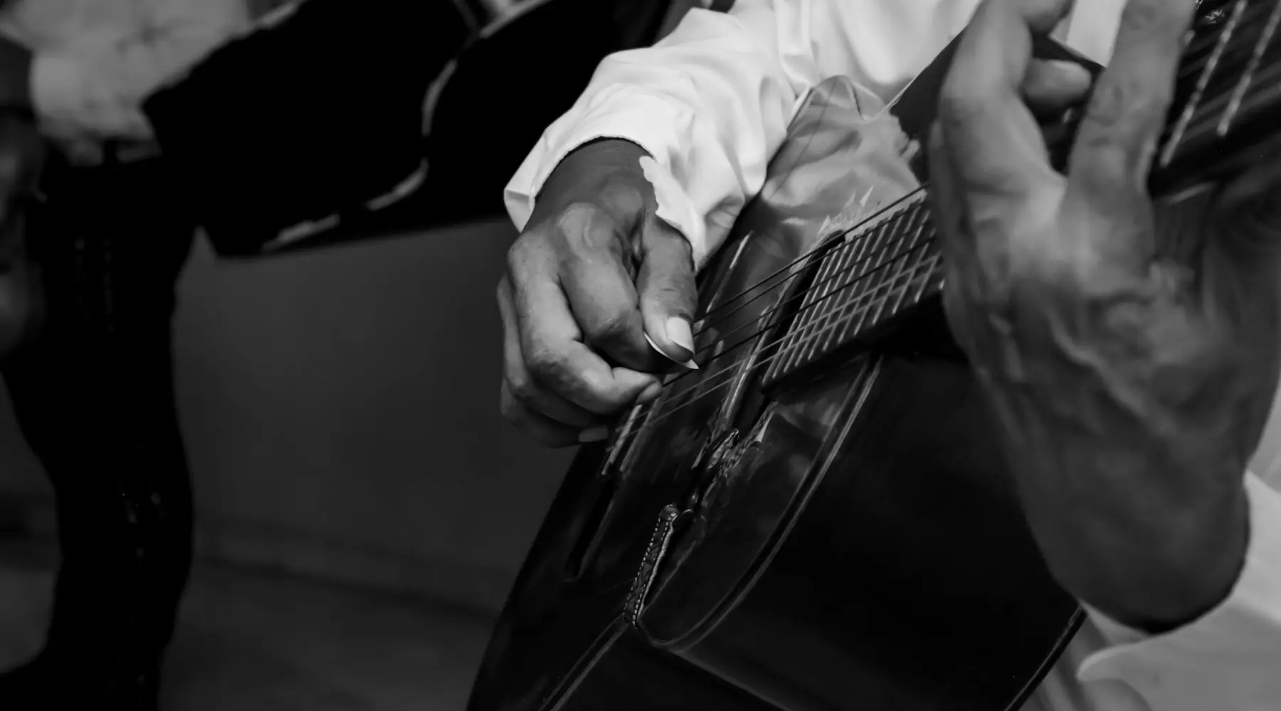 A flamenco guitarist's hands on a black guitar, shown in black and white, with the dark trousers of another musician beside him.