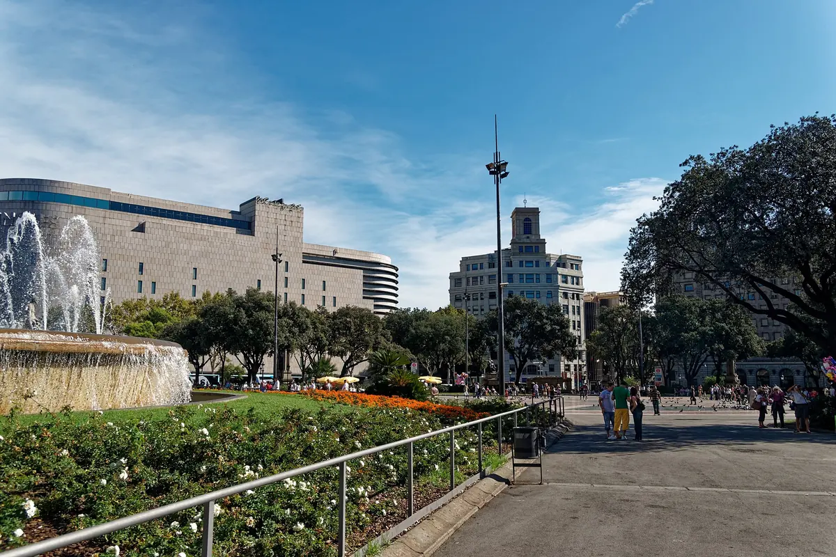 A fountain and flower beds on Plaça de Catalunya in Barcelona, with a tower and trees beyond.