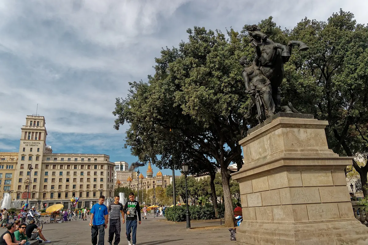 A bronze statue on a stone plinth beneath trees on Plaça de Catalunya, with people walking past and a stone tower behind.