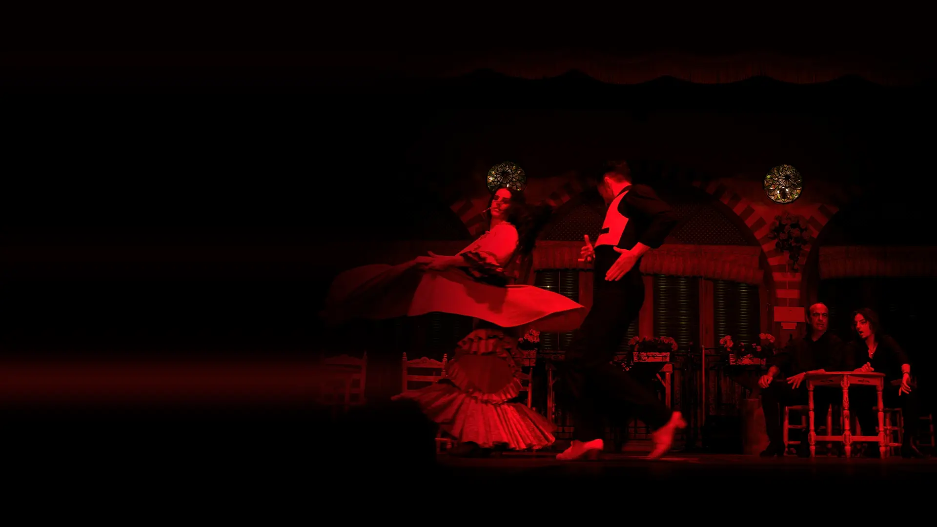A flamenco dancer in a red skirt turning beside a dancer in a black waistcoat on a stage with an arched Andalusian backdrop, with two seated musicians at the right.